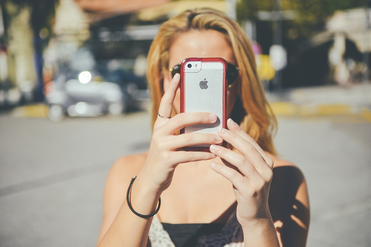 A woman facing the camera. Her face is covered by a Apple iPhone that she is holding up over her face. It looks as if she is taking a photo of the person behind the camer