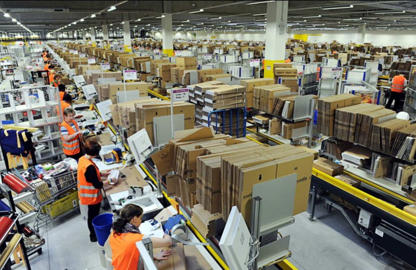 An image of the factory floor. There are a line of workers in orange vests in a production line. The perspective makes it look like the factory goes on forever