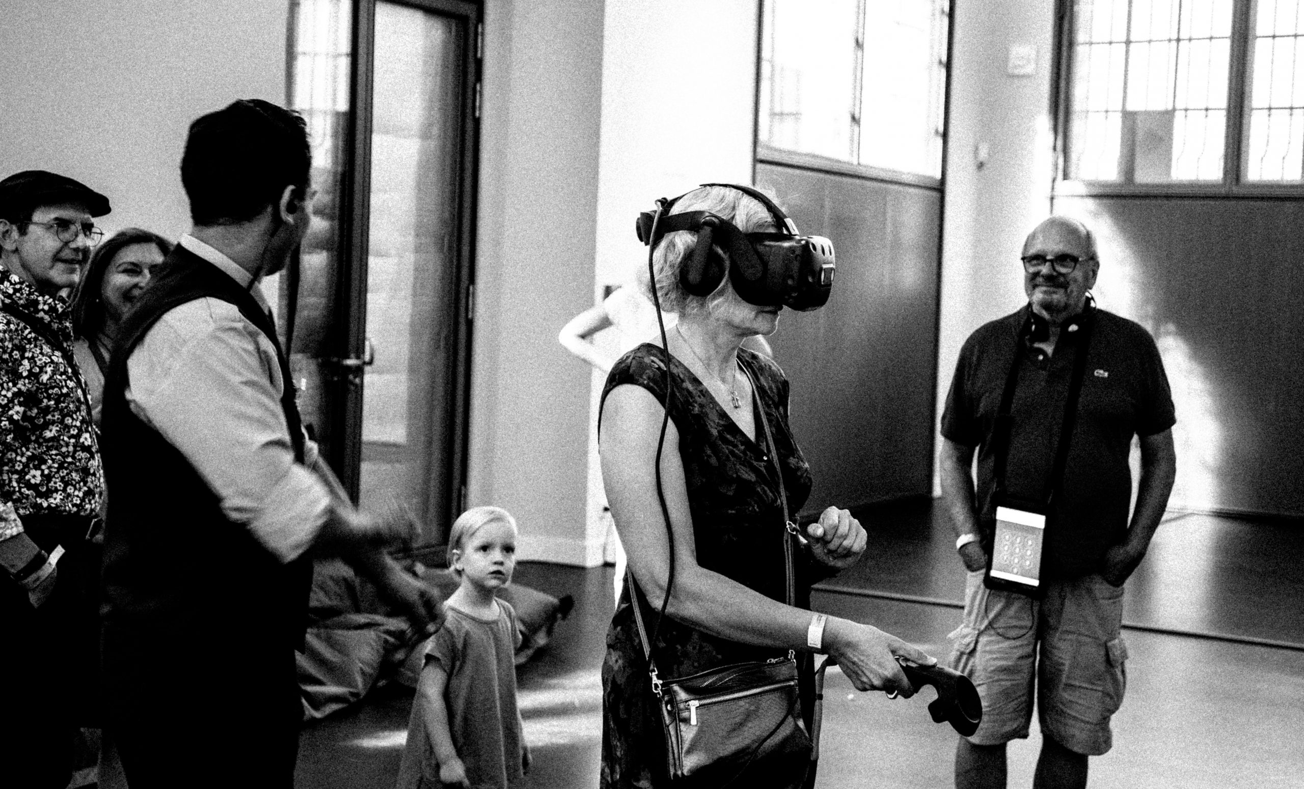 A black and white image of a woman standing in a VR headset. There are several men around her watching her