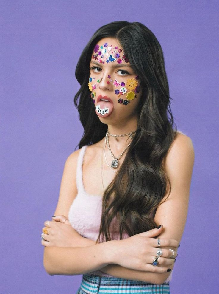 Olivia Rodrigo standing with her arms crossed and tongue out looking at the camera. She has stickers all over her face and tongue. She looks slightly annoyed
