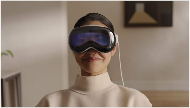 Woman sitting wearing Apple's Vision Pro headset looking directly at the camera with a slight smile. The headset covers her eyes, but on the headset is a live video feed of where her eyes are looking