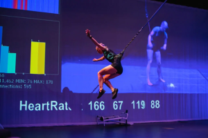 Image of the artist suspended in the air by a rope and harness system. In the background is a screen with numbers and graphs representing biometric data