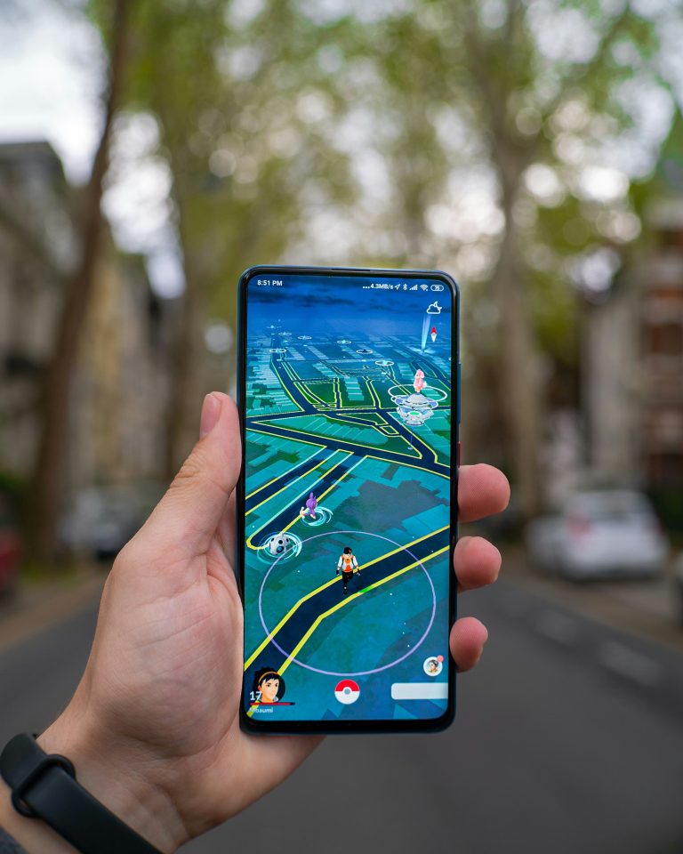 A close up of a person holding a phone up to the camera. The background is a residential street and the phone is open to the street view of pokemon go