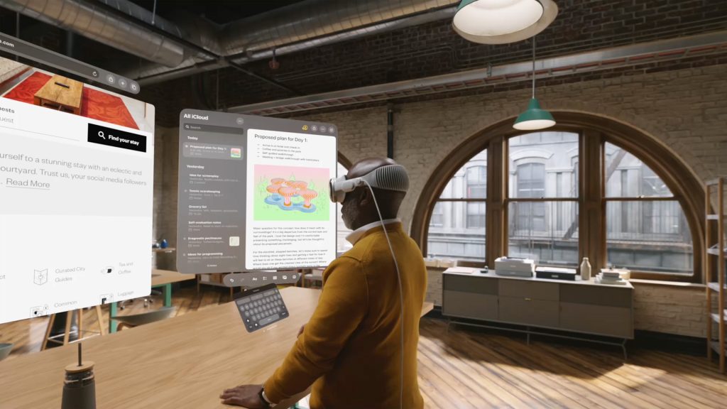 A man in a yellow sweater wearing a white Apple Vision Pro headset, standing in the middle of a room. In front of him are two augmented reality windows showing a web browser (showing a hotel booking home page) and an iCloud notes folder (the open page reading: "Proposed plan for Day 1").